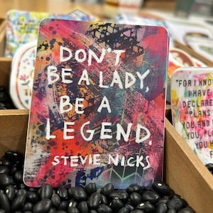 Op de afbeelding: Een kleurrijke sticker met een citaat van Stevie Nicks dat luidt: "Wees geen dame, wees een legende." De achtergrond is een mix van roze, rode, blauwe en gele verfspatten.