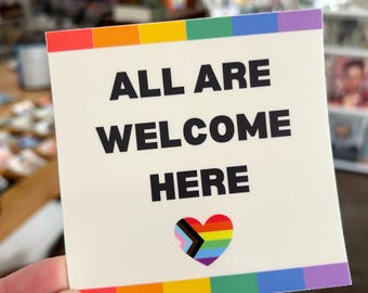 May include: Square sign with the words "ALL ARE WELCOME HERE" in black. It features a rainbow border and a heart with a rainbow and progress pride flag design. The background is blurred.