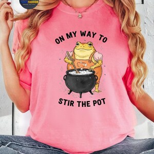 May include: Pink t-shirt with the text "ON MY WAY TO STIR THE POT" and a cartoon frog wearing overalls, stirring a cauldron. The frog is yellow and orange, and the cauldron is black. The shirt is a solid pink color.