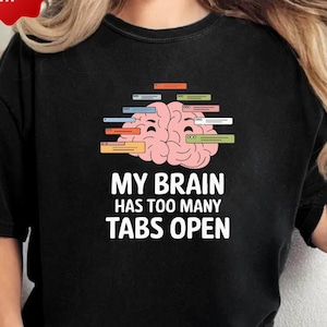 May include: Black t-shirt featuring the text "MY BRAIN HAS TOO MANY TABS OPEN" under a pink brain graphic with colourful tabs. The shirt has a crew neck and short sleeves.