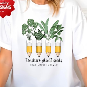 May include: White t-shirt featuring a graphic of plants in pencil-shaped pots. The text "Teachers plant seeds that grow forever" is printed below the pencils. The shirt has a crew neck and short sleeves.