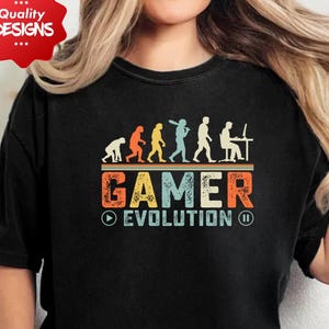 Peut inclure: T-shirt noir avec un motif coloré "Gamer Evolution". Le design présente une progression de figures humaines, du singe au joueur, au-dessus du mot "GAMER" en orange, jaune et bleu. Le t-shirt a un col rond et des manches courtes.