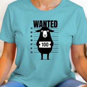 May include: A light blue t-shirt featuring a black sheep illustration with the word "WANTED" above it. The sheep holds a sign with the number "100" on it, against a height chart background.
