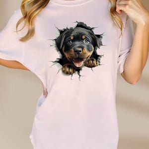 May include: A pink t-shirt with a graphic of a Rottweiler puppy peeking out of a cracked wall.