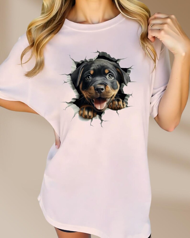 May include: A pink t-shirt with a graphic of a Rottweiler puppy peeking out of a cracked wall.