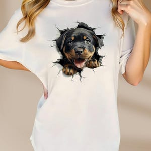 May include: White t-shirt with a graphic of a Rottweiler puppy peeking out of a cracked wall.