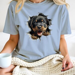 May include: A light blue t-shirt with a graphic of a Rottweiler puppy peeking out of a ripped hole. The puppy has black fur and brown eyes, and is smiling with its tongue sticking out.