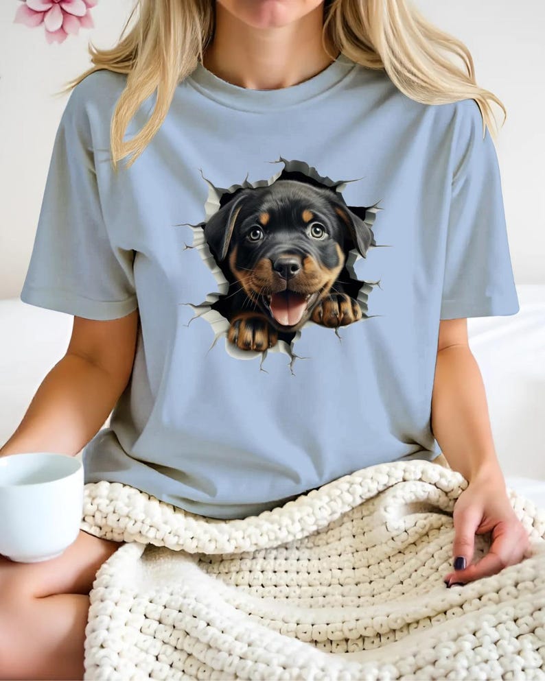 May include: A light blue t-shirt with a graphic of a Rottweiler puppy peeking out of a ripped hole. The puppy has black fur and brown eyes, and is smiling with its tongue sticking out.