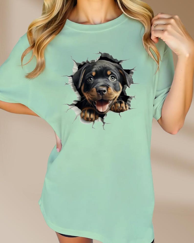 May include: A light green t-shirt with a graphic of a Rottweiler puppy peeking out of a ripped hole. The puppy is black and brown with a playful expression.
