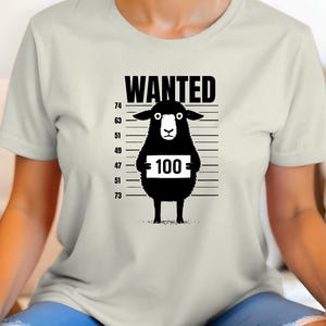 May include: Cream-colored t-shirt featuring a black graphic of a sheep in a police lineup. The word "WANTED" is above the sheep, and the sheep is holding a sign with the number "100". The shirt has a crew neck and short sleeves.