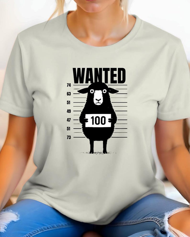 May include: Cream-colored t-shirt featuring a black graphic of a sheep in a police lineup. The word "WANTED" is above the sheep, and the sheep is holding a sign with the number "100". The shirt has a crew neck and short sleeves.