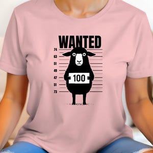 May include: A light pink t-shirt featuring a black cartoon sheep in a police lineup. The sheep holds a sign with the number 100. The word "WANTED" is printed above the sheep.