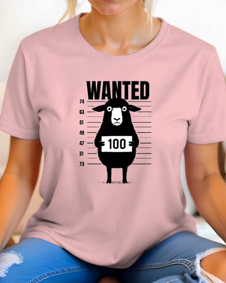 May include: A light pink t-shirt featuring a black cartoon sheep in a police lineup. The sheep holds a sign with the number 100. The word "WANTED" is printed above the sheep.
