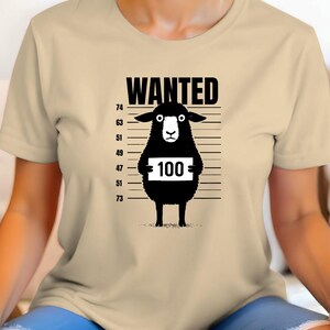 May include: A beige t-shirt featuring a black graphic of a sheep with the word "WANTED" above it. The sheep holds a sign with the number "100" on it, set against a height chart.