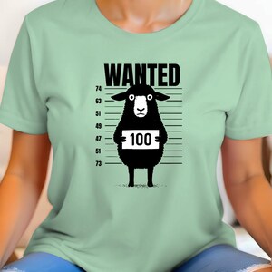 May include: A light green t-shirt featuring a black sheep illustration with the word "WANTED" above it. The sheep holds a sign with the number "100" on it, against a police lineup background.