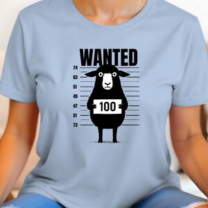 May include: Light blue t-shirt featuring a black graphic of a sheep in a police lineup. The sheep holds a sign with the number 100. The word "WANTED" is printed above the sheep.
