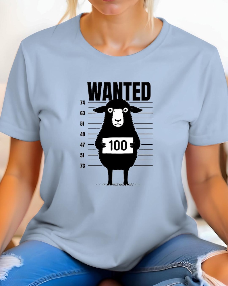 May include: Light blue t-shirt featuring a black graphic of a sheep in a police lineup. The sheep holds a sign with the number 100. The word "WANTED" is printed above the sheep.
