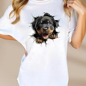 May include: White t-shirt with a graphic of a Rottweiler puppy peeking out of a cracked wall.