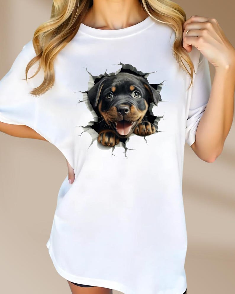 May include: White t-shirt with a graphic of a Rottweiler puppy peeking out of a cracked wall.