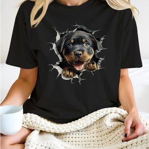 May include: Black t-shirt with a graphic of a Rottweiler puppy peeking out of a ripped hole in the fabric.