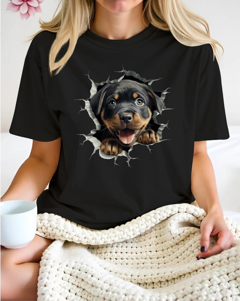 May include: Black t-shirt with a graphic of a Rottweiler puppy peeking out of a ripped hole in the fabric.