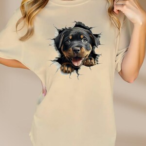 May include: A beige t-shirt with a graphic of a Rottweiler puppy peeking out of a cracked wall.