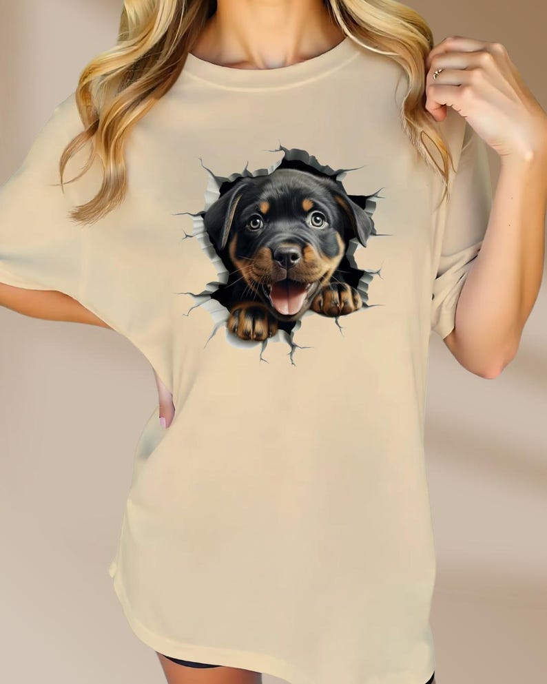 May include: A beige t-shirt with a graphic of a Rottweiler puppy peeking out of a cracked wall.