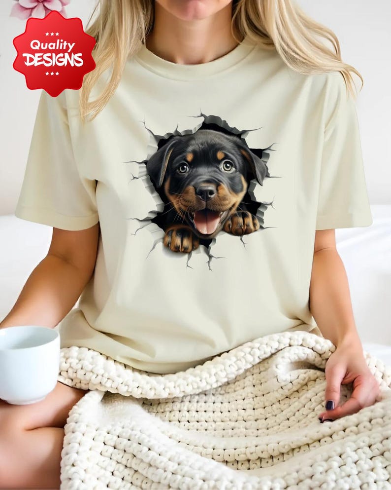 May include: A beige t-shirt with a graphic of a Rottweiler puppy peeking out of a cracked wall. The puppy has black fur with brown markings and is looking directly at the viewer.