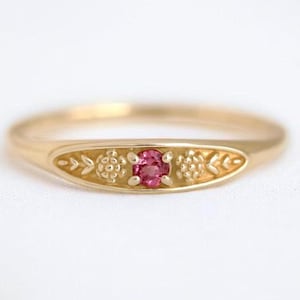 以下が含まれることがあります： 小さな丸いピンク色の宝石を特徴とする、中央の楕円形のデザインのゴールドリング。リングには、宝石の両側に花柄と葉柄があります。リングは白い背景にセットされています。