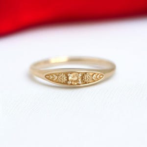 Puede incluir: Un anillo de oro con una piedra central cuadrada, flanqueada por diseños florales y de hojas. El anillo tiene una banda lisa y pulida y una pieza central decorativa. El anillo está sobre un fondo blanco con un acento rojo.