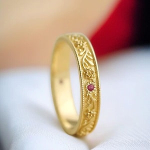May include: A gold ring with an intricate floral pattern and a small, round, red gemstone. The ring's band features detailed carvings of leaves and flowers. The ring is set against a soft, blurred background, highlighting its ornate details.
