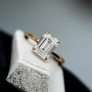 May include: A gold ring with an emerald-cut diamond. The clear, rectangular diamond is held by four prongs. The band is thin, polished gold. The ring is shown on a textured, white surface against a blurred background.