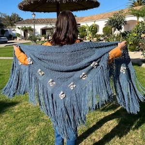 May include: A blue crocheted shawl with long fringe and floral embellishments. The shawl is held open, showcasing its intricate design and texture. The background features a sunny outdoor setting with a thatched umbrella and a building.