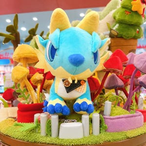 Puede incluir: Un peluche de criatura azul y amarilla con forma de drag&oacute;n, con ojos grandes y una expresi&oacute;n amigable. Tiene cuernos y alas amarillas, y est&aacute; rodeado de coloridas decoraciones de setas y plantas. El peluche est&aacute; sobre una exhibici&oacute;n con una valla blanca.