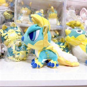 Puede incluir: Un peluche azul y amarillo con forma de criatura con ojos grandes, una larga cola y patas con garras. El peluche tiene una cola blanca y esponjosa y detalles amarillos en la cabeza y el cuerpo. Otros peluches similares son visibles en el fondo.