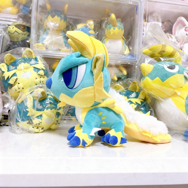 Puede incluir: Un peluche azul y amarillo con forma de criatura con ojos grandes, una larga cola y patas con garras. El peluche tiene una cola blanca y esponjosa y detalles amarillos en la cabeza y el cuerpo. Otros peluches similares son visibles en el fondo.