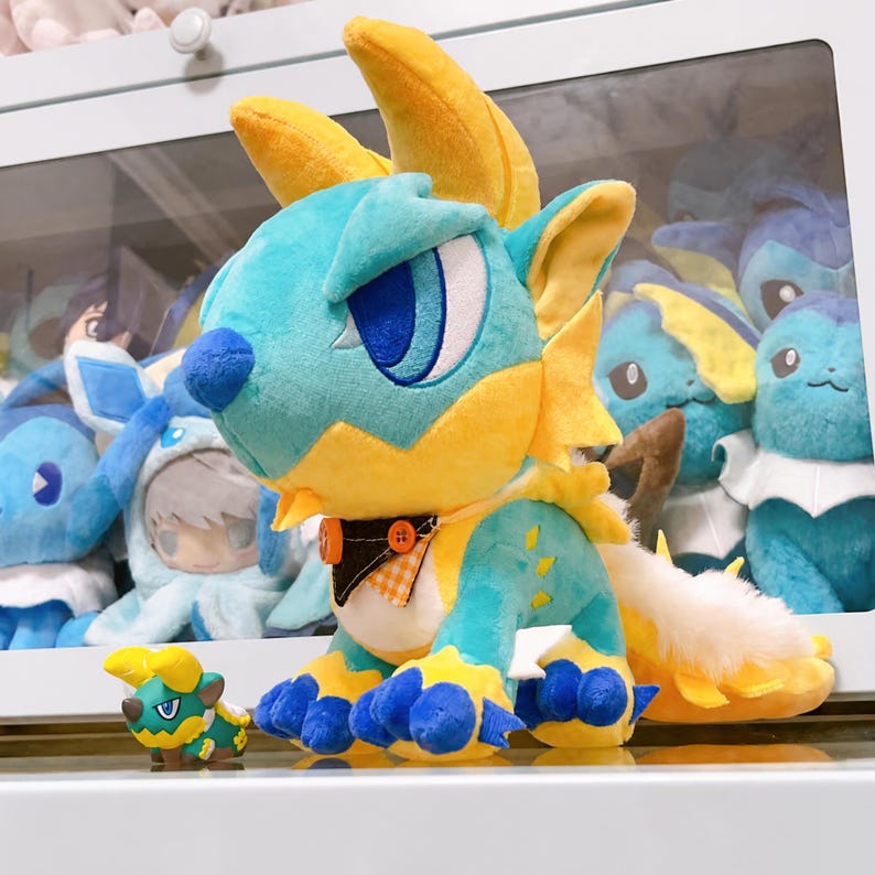 Puede incluir: Un gran peluche de drag&oacute;n en tonos azules, amarillos y naranjas, con una figura m&aacute;s peque&ntilde;a. El peluche tiene ojos grandes, cuernos y una cola esponjosa. Otros peluches similares son visibles en el fondo.