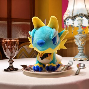 Puede incluir: Un peluche de drag&oacute;n en tonos azules y amarillos est&aacute; sobre un plato decorativo. El drag&oacute;n tiene ojos grandes, cuernos y una cola esponjosa. Est&aacute; colocado en una mesa con cubiertos y una copa de vino, creando una escena caprichosa.