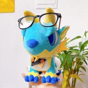 Puede incluir: Un drag&oacute;n de peluche en tonos azules y amarillos, con gafas de montura negra. El drag&oacute;n tiene un hocico azul, ojos grandes azules y una peque&ntilde;a pajarita. El juguete es suave y tierno, con un dise&ntilde;o caprichoso.
