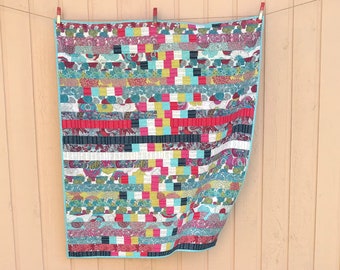 May include: A colorful quilt with a patchwork design featuring various shades of red, blue, green, yellow, and black. The quilt is hanging on a light pink wooden wall.