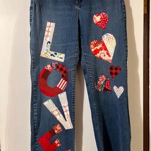 May include: Blue denim jeans with patchwork hearts and the word "LOVE" appliquéd on the front. The letters and hearts are made from various patterned fabrics in red, white, and other colors. A unique, handmade clothing item.