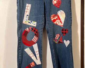May include: Blue denim jeans with patchwork hearts and the word "LOVE" appliquéd on the front. The letters and hearts are made from various patterned fabrics in red, white, and other colors. A unique, handmade clothing item.