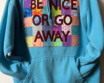 May include: A bright blue hooded sweatshirt featuring a large, rectangular patchwork panel on the chest. The panel consists of various colorful patterned fabric squares. Dark maroon text reads BE NICE OR GO AWAY across the center of the patch.