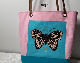 May include: Pink and teal tote bag with brown leather handles. The bag features a large butterfly graphic on a teal panel, with the text "Bag 1" in the upper right corner. The bag has a teal bottom and pink sides.