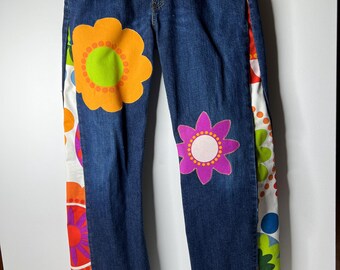 May include: Dark blue denim jeans with large, colorful flower appliqués. The jeans feature an orange flower with a green center and a pink flower with a white center. The sides have a patterned fabric with more flowers.