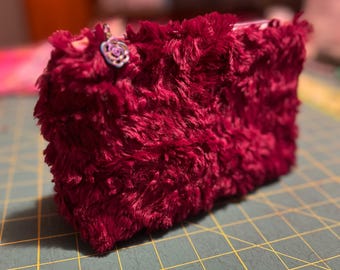 May include: A rectangular, plush burgundy-colored pouch with a fluffy texture. A small, decorative charm with a floral design hangs from the top. The pouch is resting on a green cutting mat with yellow grid lines.