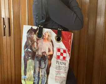 May include: A repurposed tote bag made from a Purina horse feed bag. The bag features a brown horse and a woman in jeans and a white shirt. The bag has black leather straps with brass grommets and a red and white Purina logo.