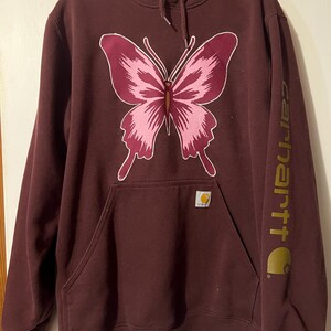 Puede incluir: Sudadera con capucha Carhartt burdeos con un gran dise&ntilde;o de mariposa rosa. La sudadera tiene una capucha con cordones, un bolsillo de canguro y el logotipo de Carhartt en la manga y el bolsillo. Una prenda casual y c&oacute;moda.