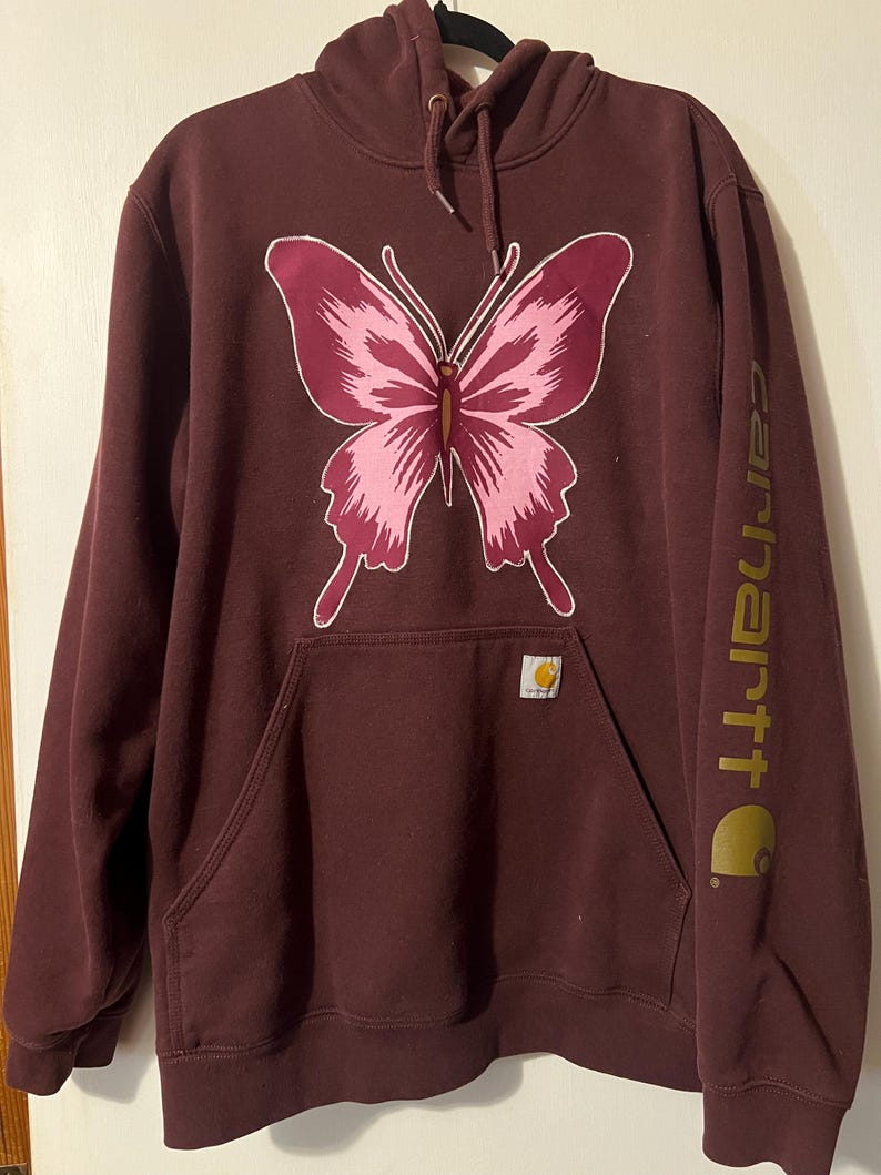 Puede incluir: Sudadera con capucha Carhartt burdeos con un gran dise&ntilde;o de mariposa rosa. La sudadera tiene una capucha con cordones, un bolsillo de canguro y el logotipo de Carhartt en la manga y el bolsillo. Una prenda casual y c&oacute;moda.