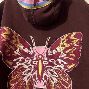 Puede incluir: Una sudadera con capucha granate con un gran dise&ntilde;o de mariposa en la espalda. La mariposa tiene detalles dorados y blancos. La capucha tiene un gr&aacute;fico de mariposa rosa y rayas azules y amarillas.